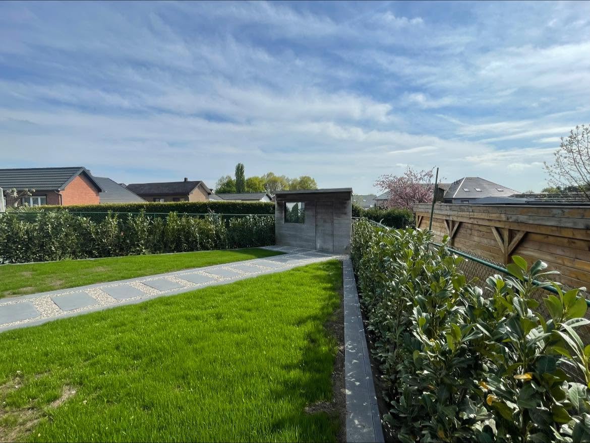 Groen gazon met een pad van tegels en grind naar een tuinhuis, omzoomd door hagen